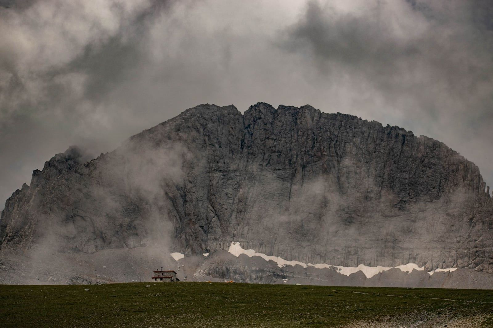 A mountain refuge sits on an alpine plateau beneath the cloud-shrouded limestone cliffs of Mount Olympus in Greece.