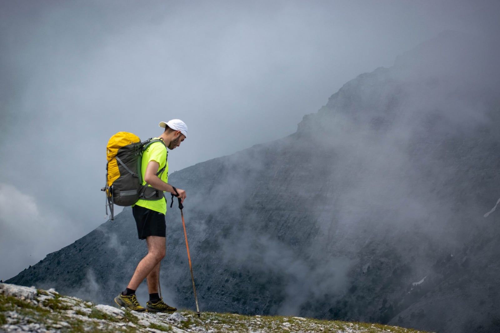 A hiker with a backpack and trekking poles walks along a rocky mountain trail in the mist on Mount Olympus.