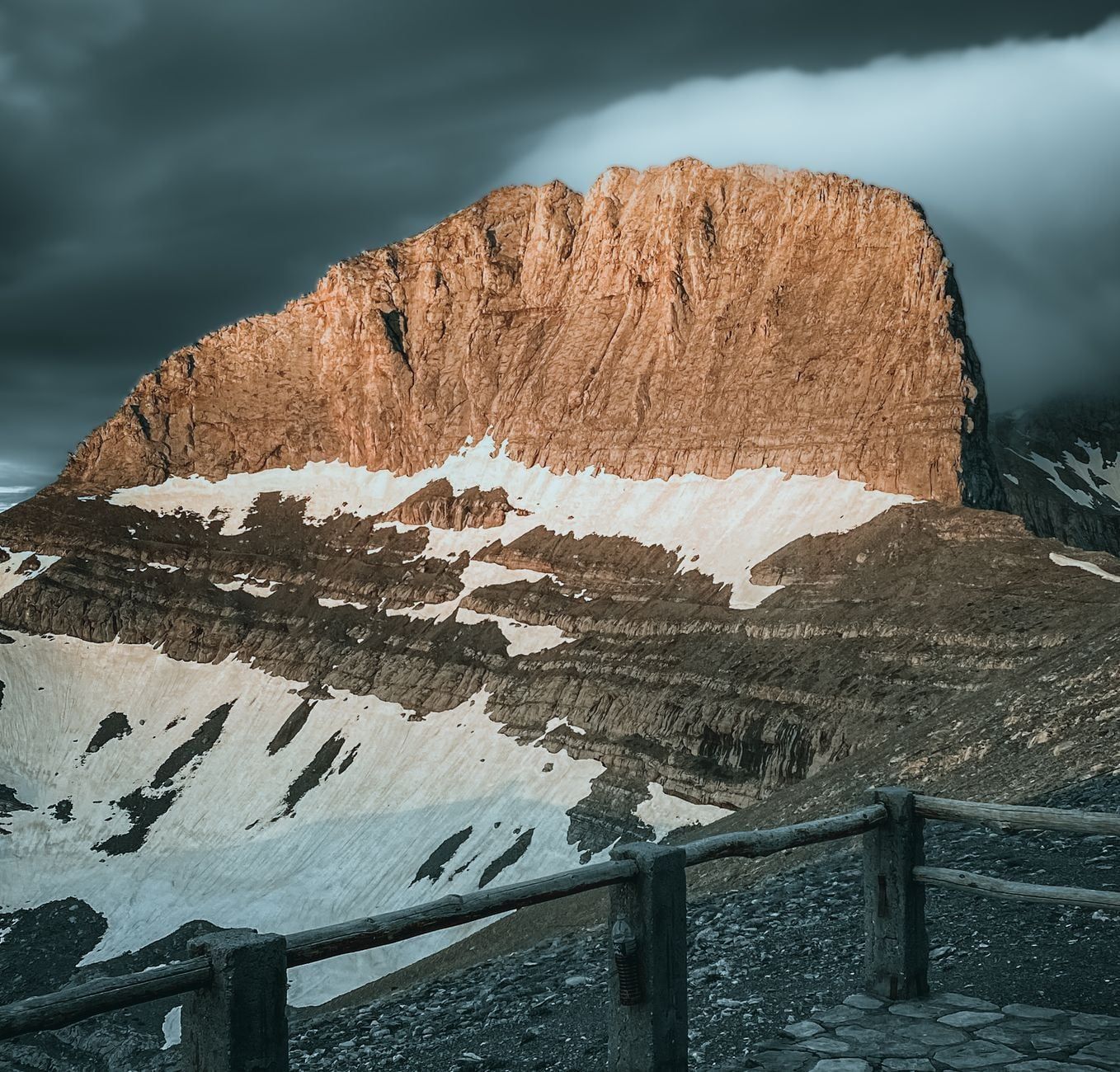 Sunlight illuminates the sheer rocky summit wall and snowfields of Mount Olympus beneath dark clouds.